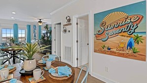 A dining area with a table set for four, a ceiling fan, and a wall with a beach-themed poster. - Sunset Serenity marsh view w/pool (North Myrtle Beach)