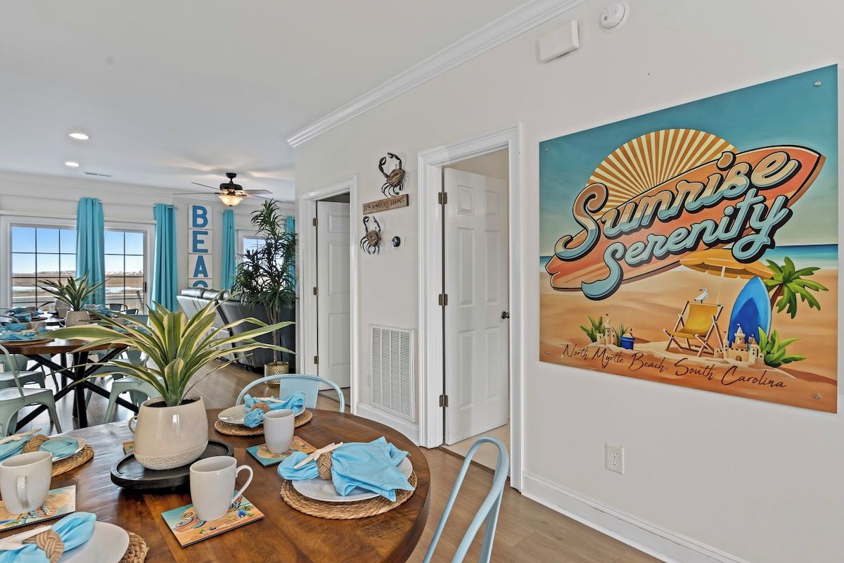 A dining area with a wooden table set for a meal, a wall with a 'Sunrise Serenity' mural, and a 'BEAUTY' sign.