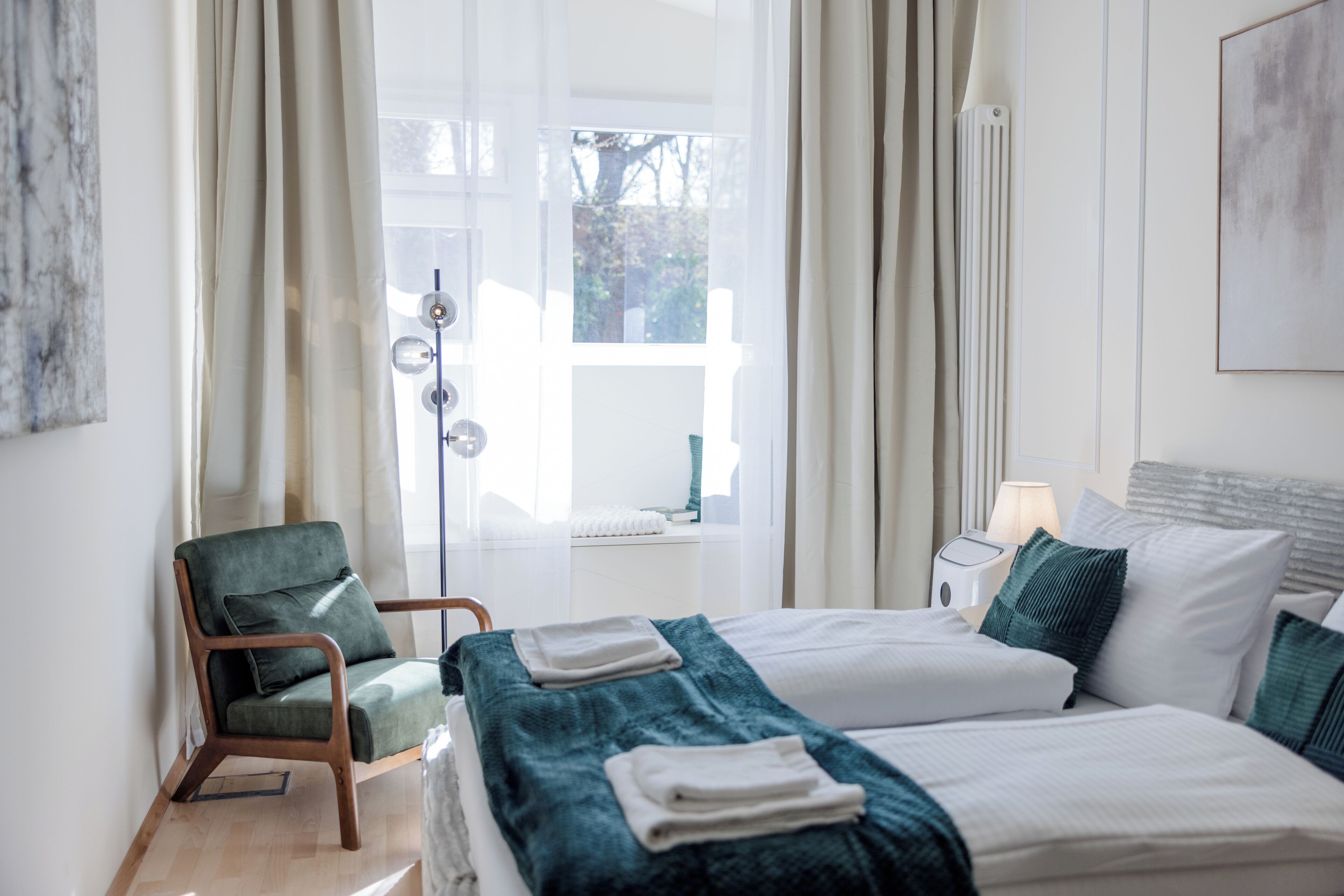 Una habitación de hotel con cama, una silla, ventana con cortinas, radiador y mesita de noche con lámpara.