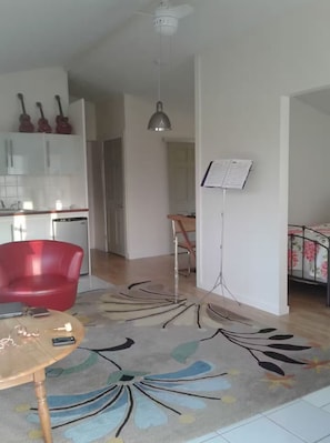 A modern living room with a red chair, a coffee table, a rug, a dining area with a chair, and a kitchen area with cabinets and a hanging light fixture.
