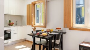 A modern kitchen with white cabinets, a dining table with black chairs, and a breakfast setup including fruits and a croissant. - 3 Stylish Flats w/ Balcony & Garage by HostWise (Porto)