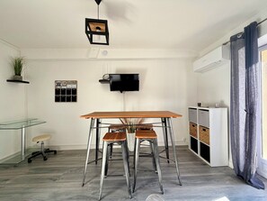 Une cuisine moderne avec une table en bois, deux tabourets de bar, un luminaire suspendu, un téléviseur fixé au mur et une plante.