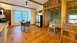 A room with wooden floors, a dining table with chairs, a sofa, a TV, and a kitchen area with a refrigerator and cabinets.
