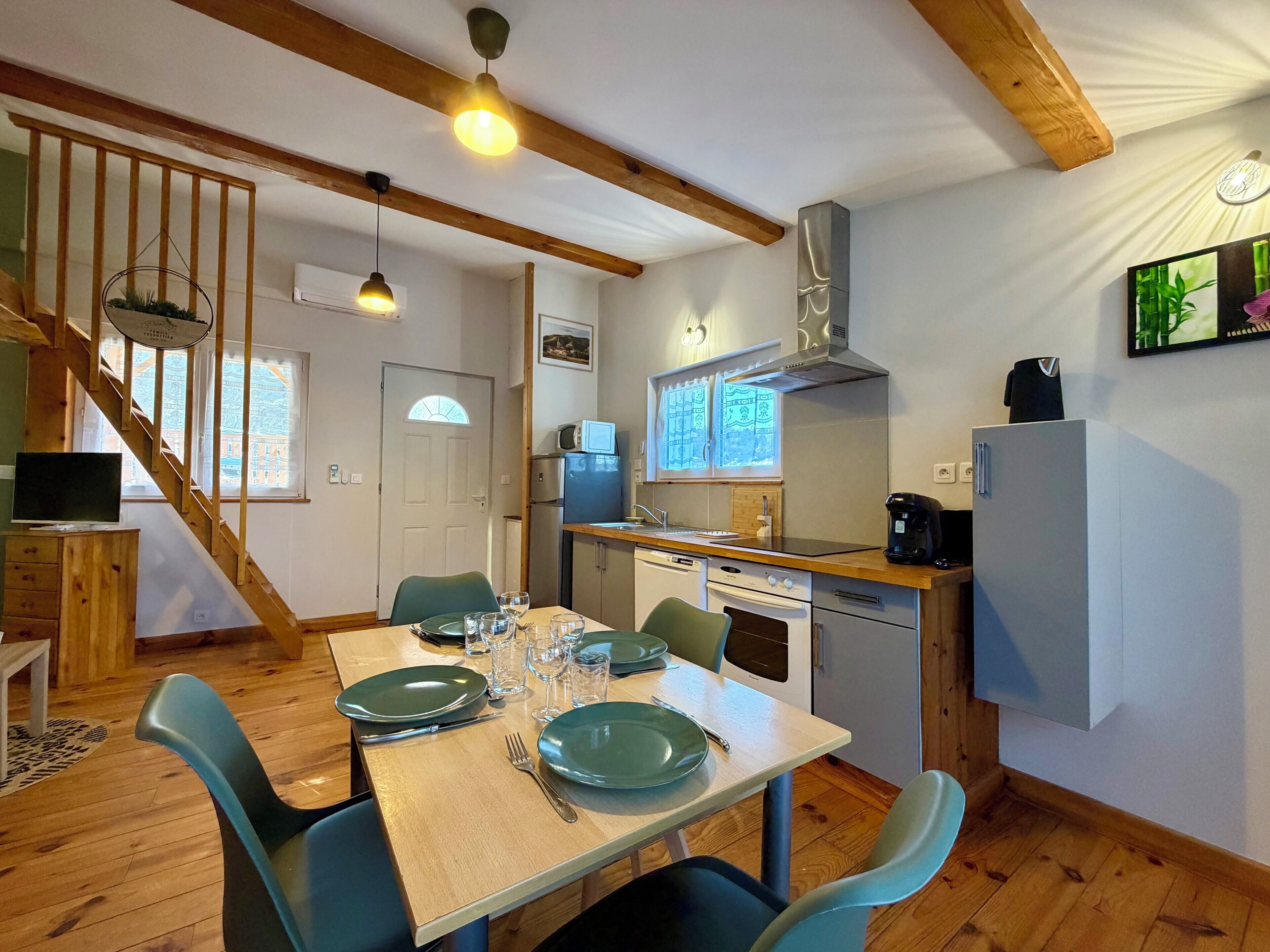 Een eetgedeelte met een houten tafel voor vier personen, een keuken met moderne apparatuur, en een trap naar een hoger gelegen verdieping.
