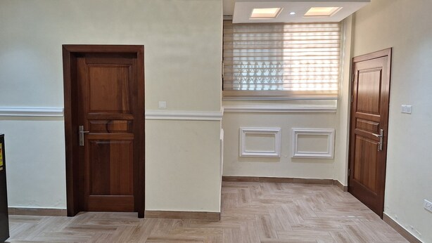 A hallway with wooden doors, a herringbone-patterned floor, recessed ceiling lights, and a window with blinds. - Cozy 1-bedroom apartment in perfect Greater Accra with AC, Fibre Internet (Greater Accra)