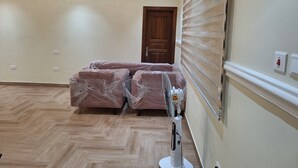 A room with a wooden floor, a bed covered in plastic, a standing fan, a door, and a window with blinds. - Cozy 1-bedroom apartment in perfect Greater Accra with AC, Fibre Internet (Greater Accra)