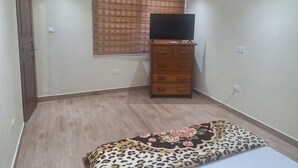 A bedroom with a bed, a TV on a wooden dresser, a window with blinds, an air conditioner, and a door. - Cozy 1-bedroom apartment in perfect Greater Accra with AC, Fibre Internet (Greater Accra)