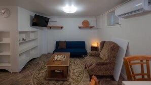 A living room with a blue sofa, a wooden coffee table with a chessboard, a patterned armchair, a TV mounted on the wall, and an air conditioning unit.
