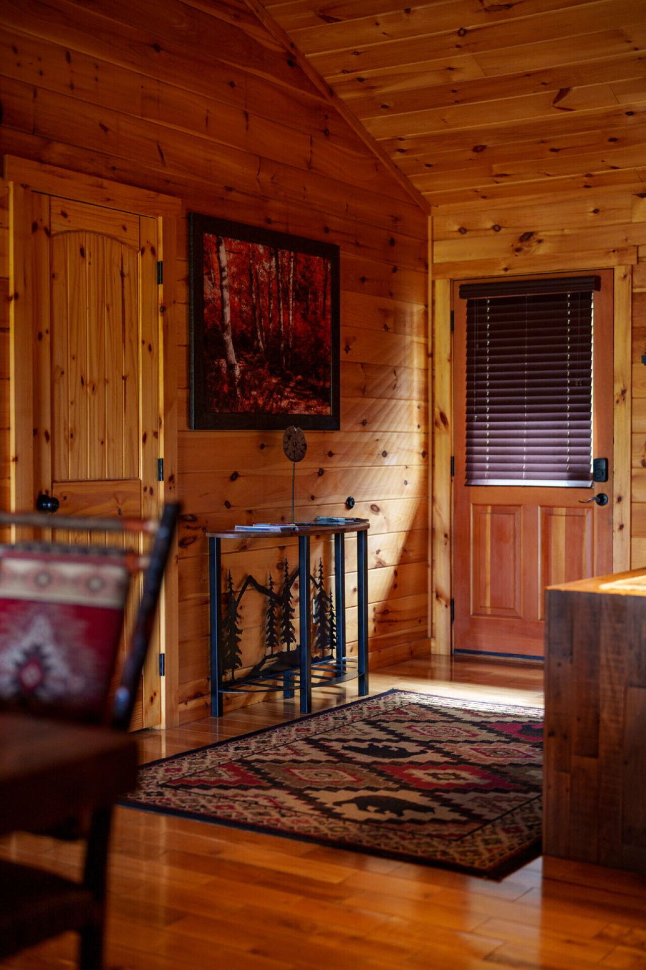 A wooden interior with a door, a framed picture, a small table, and a patterned rug.