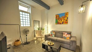Una sala de estar con un sofá gris, una mesa de centro negra, un ventilador de pie, una ventana y un cuadro en la pared.