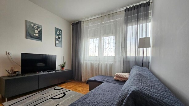 A modern bedroom with a TV, two beds, a sofa, a window with curtains, and artwork on the wall. - Apartman Fitt (Zagreb)