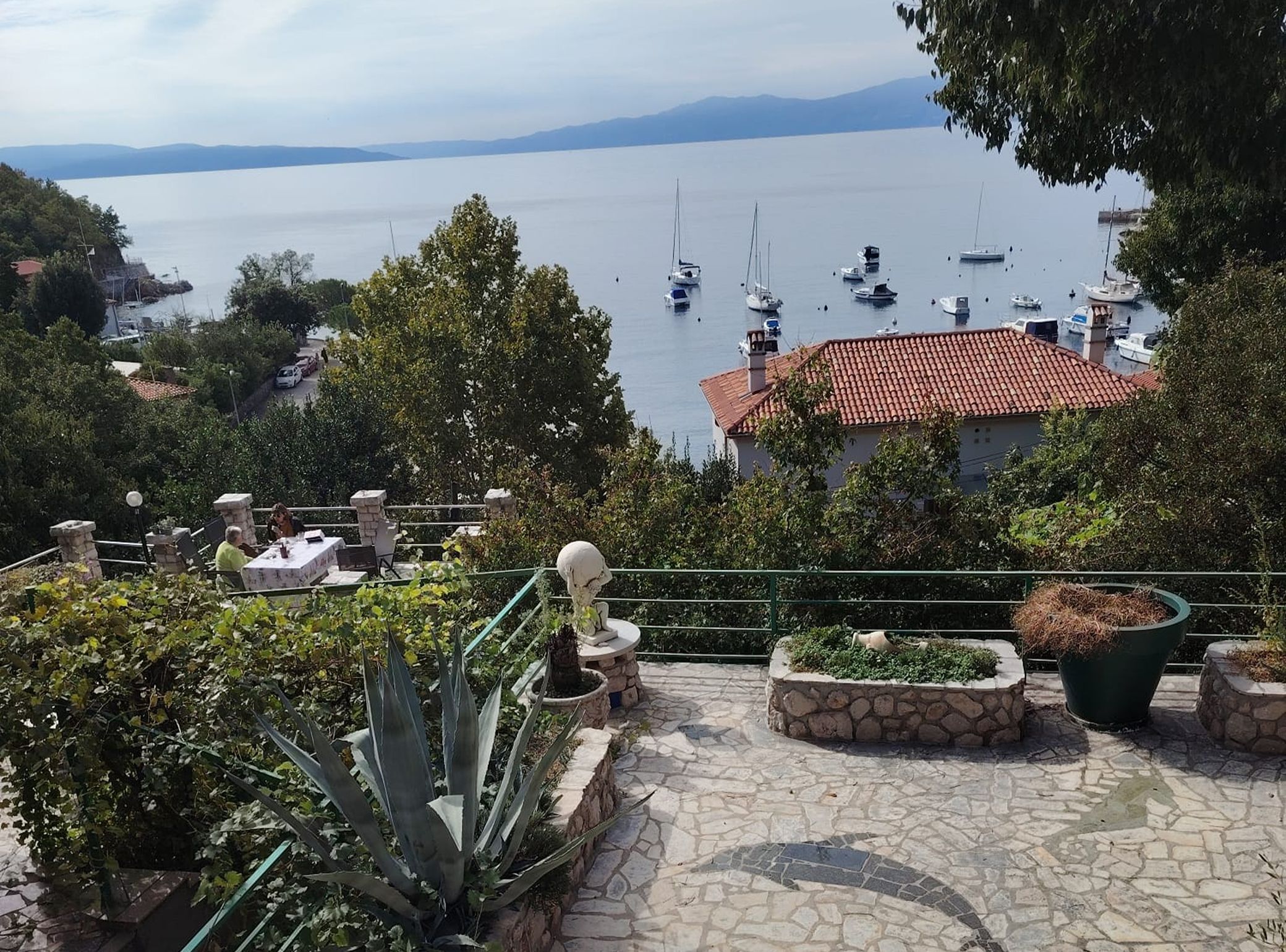 Vista costera con un sendero de piedra, plantas en macetas y una mesa con sillas.