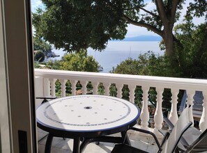 A balcony with a round table and chairs, overlooking a coastal view with trees and a distant shoreline. - Comfort Kostrena Apartments Rijeka (Kostrena)
