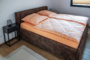 Ett sovrum med en brun stoppad säng, en grå matta, ett sängbord med en lampa och ett fönster med persienner.