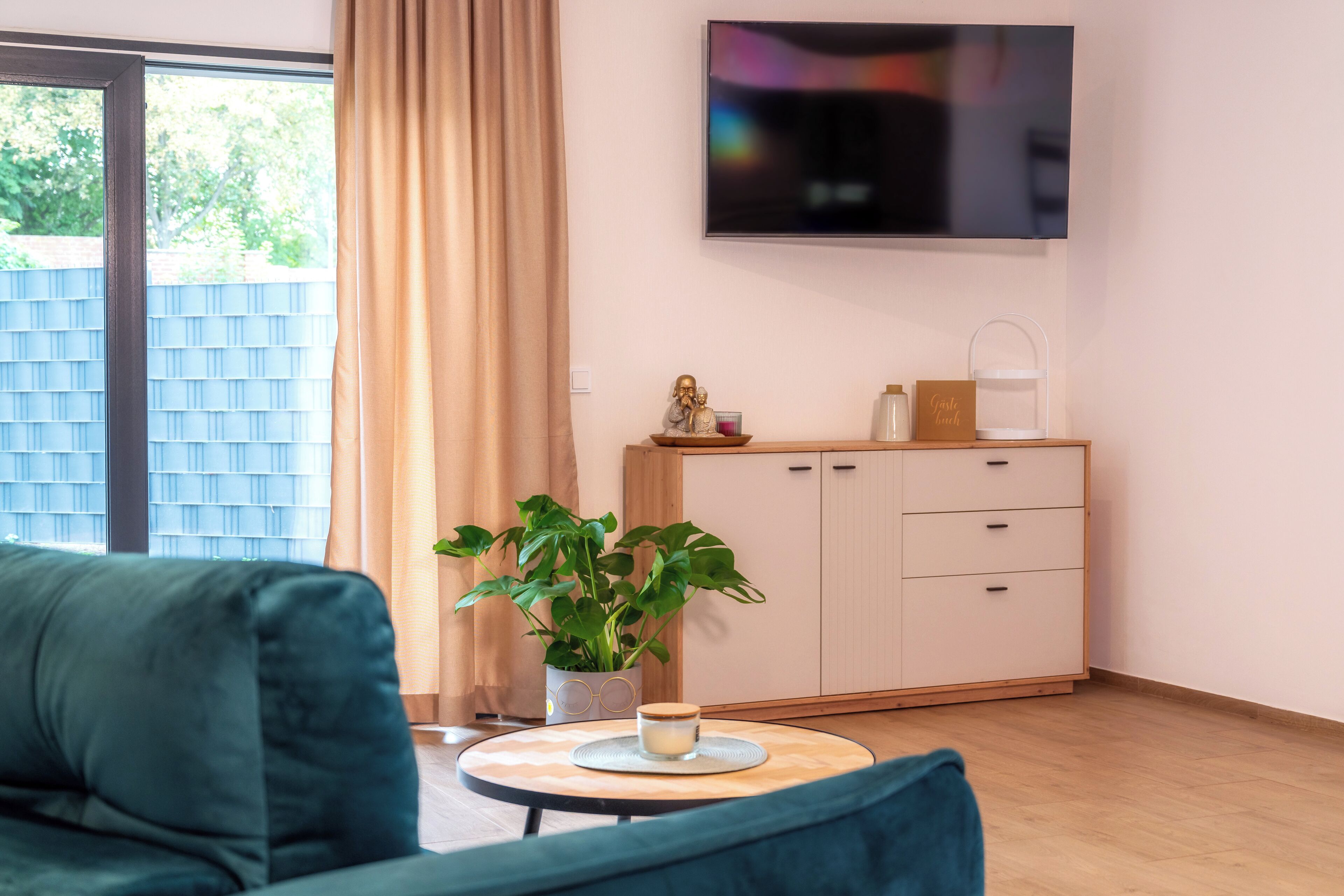 Ein modernes Wohnzimmer mit einer Couch, einem Couchtisch, einer Zimmerpflanze, einem an der Wand montierten Fernseher und einer Kommode mit Schubladen.