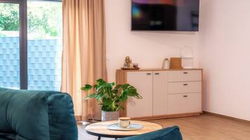 Ein modernes Wohnzimmer mit einer Couch, einem Couchtisch, einer Zimmerpflanze, einem an der Wand montierten Fernseher und einer Kommode mit Schubladen.
