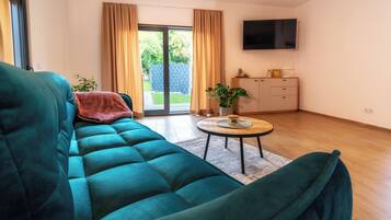 Un salon avec un canapé de couleur turquoise, une petite table ronde ornée d’une plante, un meuble et un téléviseur fixé au mur.