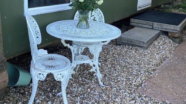 A small outdoor seating area with a table and two chairs, a vase with flowers, and a building with a door and window. - Wimbleball Lake View Cabin Sleeps up to 3 (Dulverton)