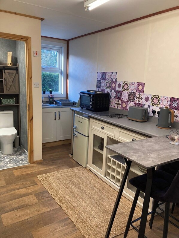 A compact kitchen with a microwave, toaster, and a small dining area with a table and chairs. - Wimbleball Lake View Cabin Sleeps up to 3 (Dulverton)