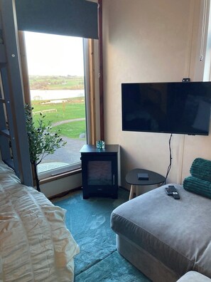 A hotel room with a bed, a television, a small table, a heater, and a view of a lake. - Wimbleball Lake View Cabin Sleeps up to 3 (Dulverton)