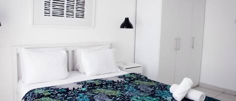 A bedroom with a bed, white headboard, a wardrobe, a nightstand, and a wall-mounted lamp.