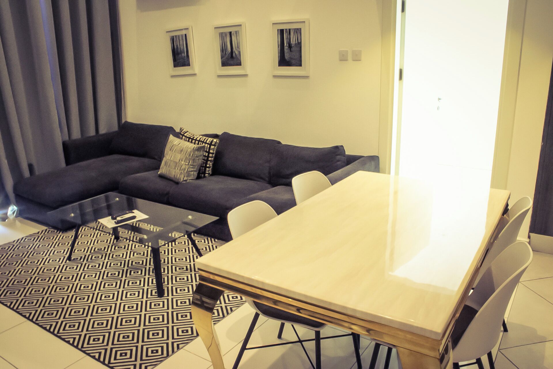 A modern living room with a dark-colored sofa, a glass coffee table, and a light-colored dining table with chairs. There are three framed pictures on the wall above the sofa.