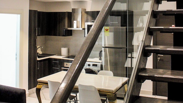A modern kitchen with dark cabinetry, a stainless steel refrigerator, and a dining area with a glass table and chairs. - Embassy Gardens Exquisite (Accra)