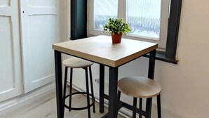 Ein Eckzimmer mit Fenster, einem Tisch, zwei Hocker und einem Blumentopf.