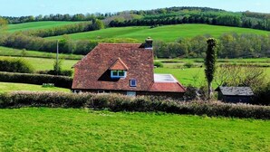 Une maison de campagne avec un toit en pente, entourée de champs verdoyants et d’un paysage vallonné.