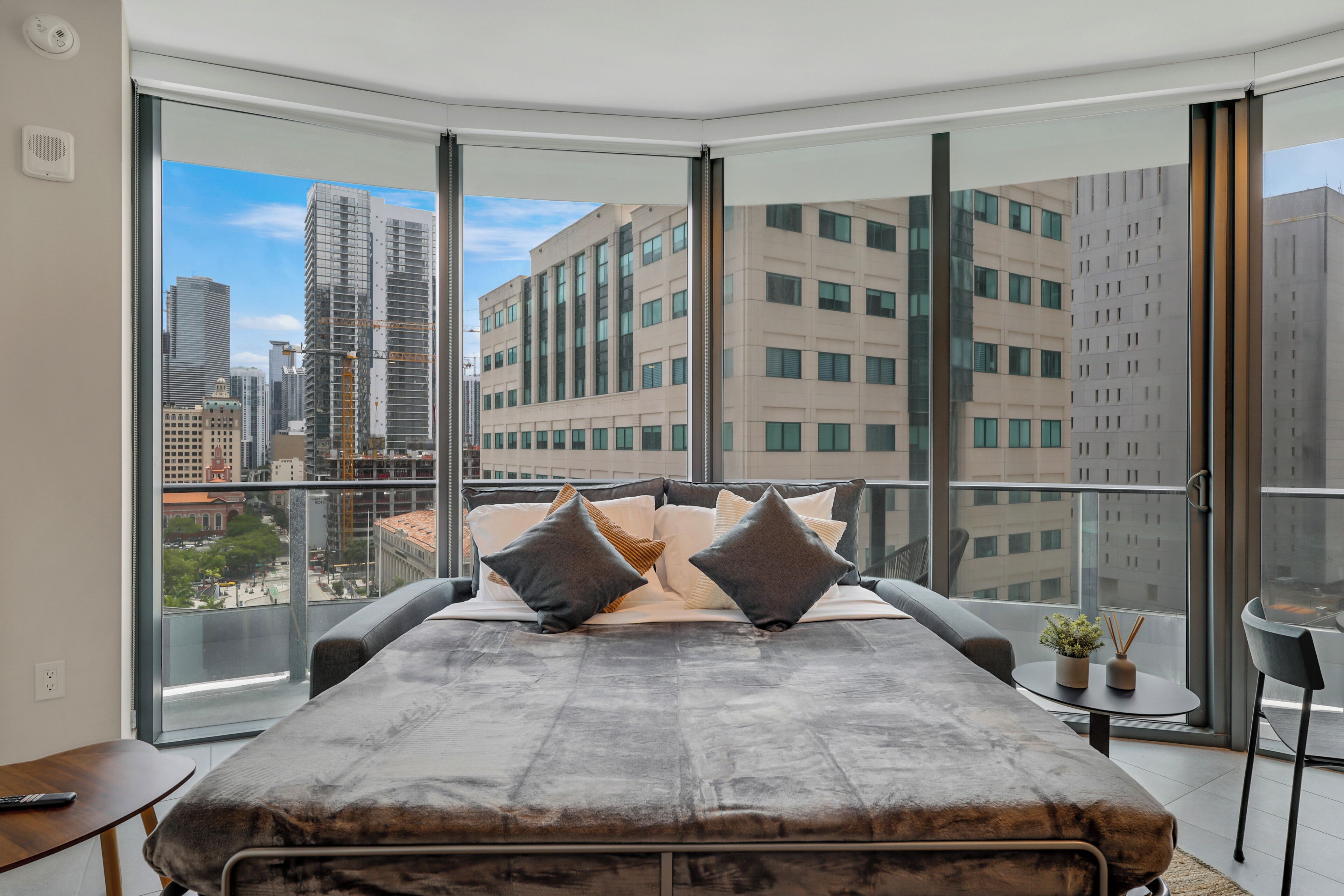A modern bedroom with a large bed, a small table, and a chair, featuring a city view through floor-to-ceiling windows.