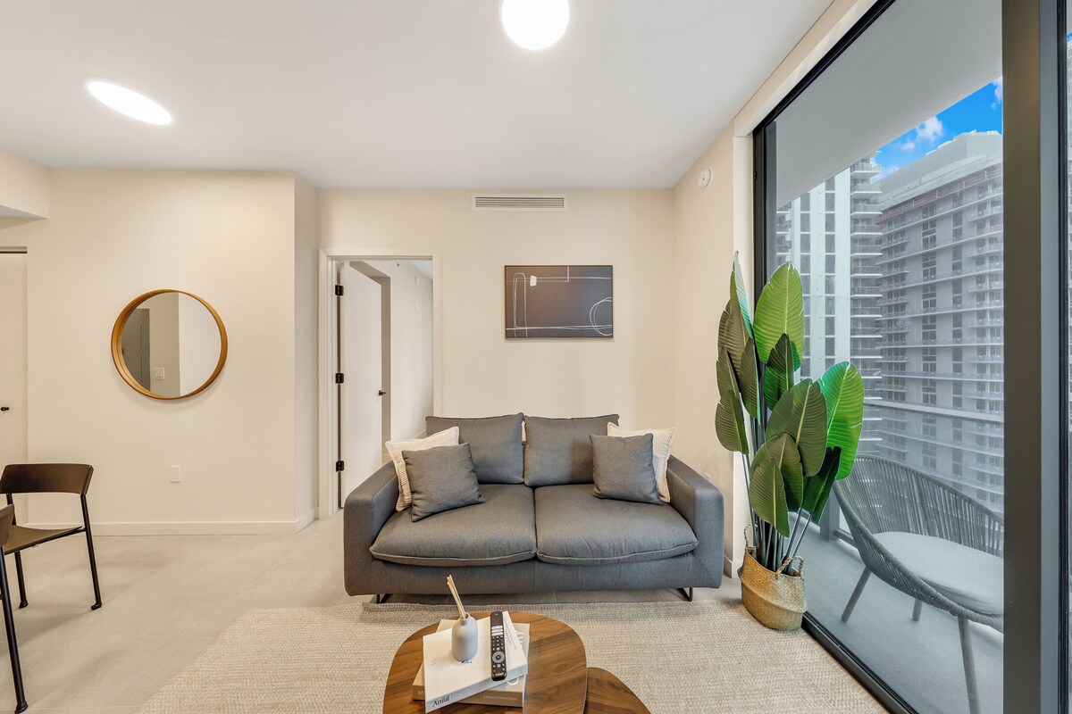 A modern living room with a grey sofa, a round mirror, a coffee table with books and a remote, and a large window with a view of a building.