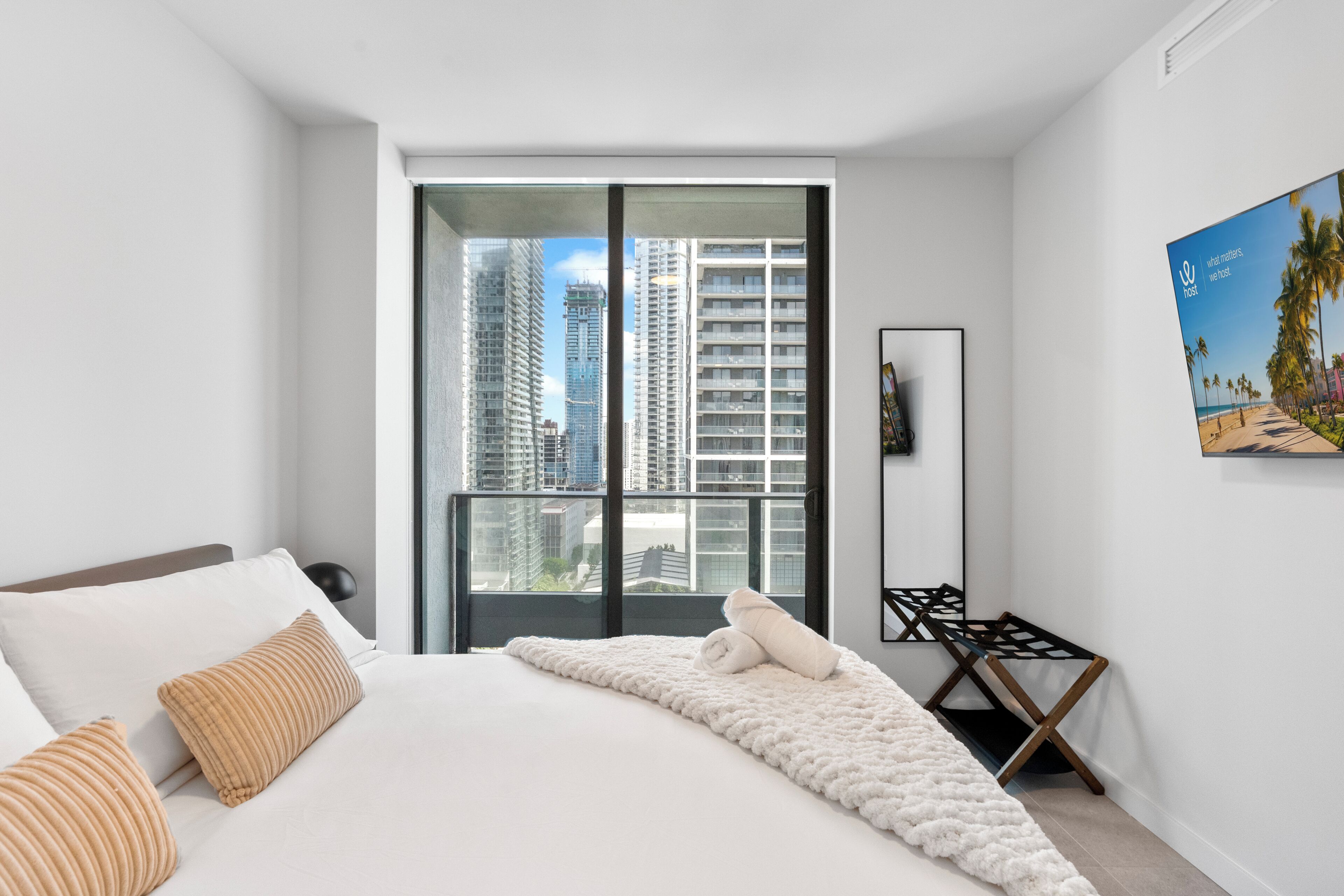 Habitación de hotel moderna con una cama grande, vistas a edificios altos, una obra de arte enmarcada y una mesita pequeña.