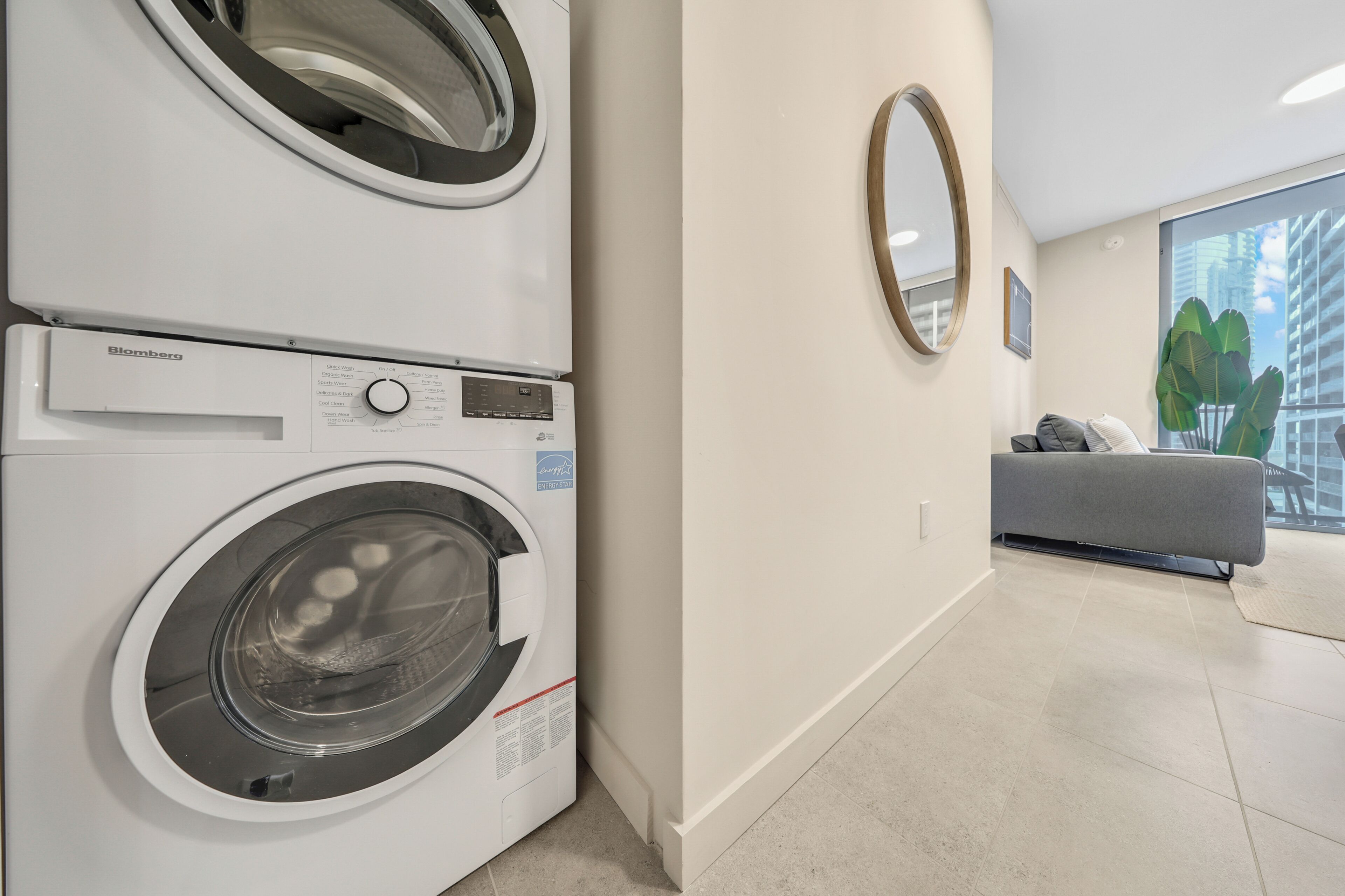 A modern laundry room with a front-loading washer and dryer, a grey sofa, and a large window with a view of buildings.