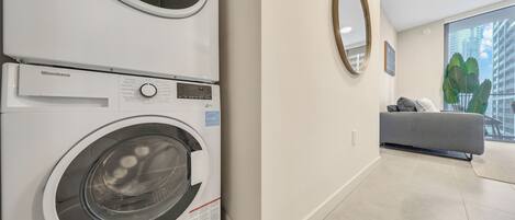 A modern laundry room with a front-loading washer and dryer, a grey sofa, and a large window with a view of buildings.