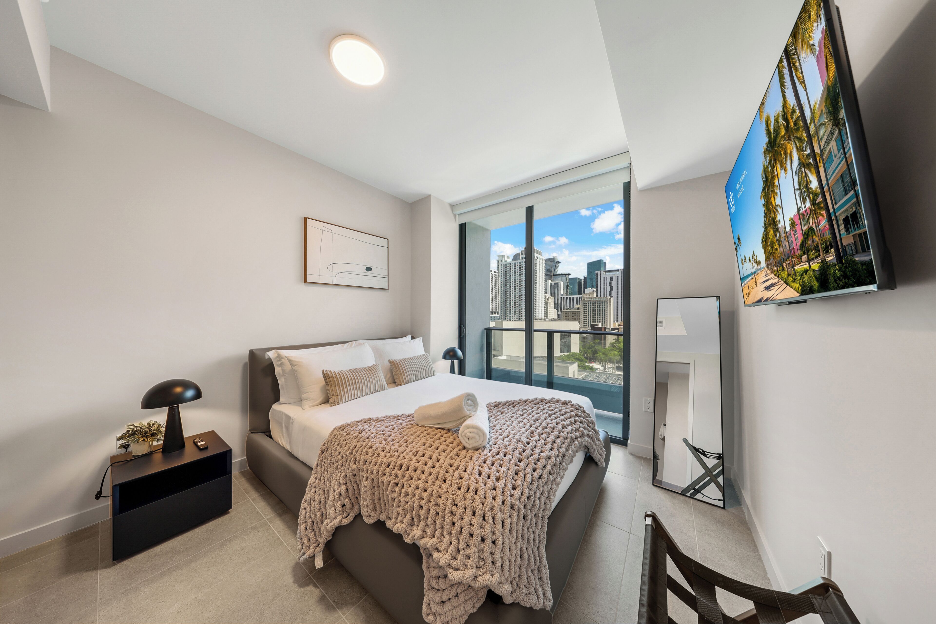 A modern bedroom with a large bed, a nightstand, a lamp, a TV, and a city view.