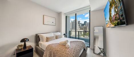 A modern bedroom with a large bed, a nightstand, a lamp, a TV, and a city view.