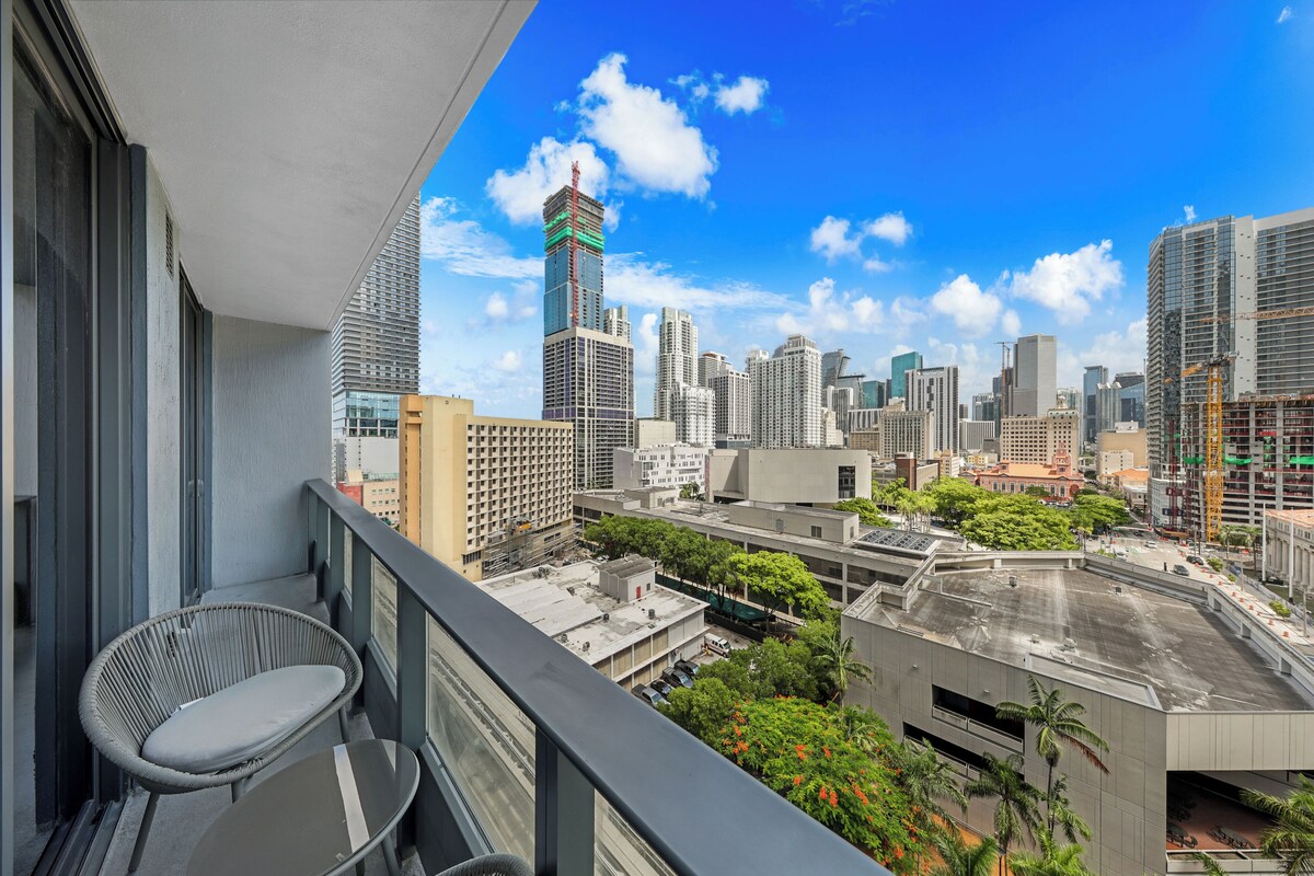 A balcony with a wicker chair and a table, overlooking a cityscape with high-rise buildings and construction cranes.