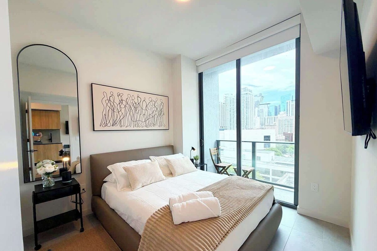 A modern hotel room with a large bed, a city view, a flat-screen TV, and a minimalist decor.