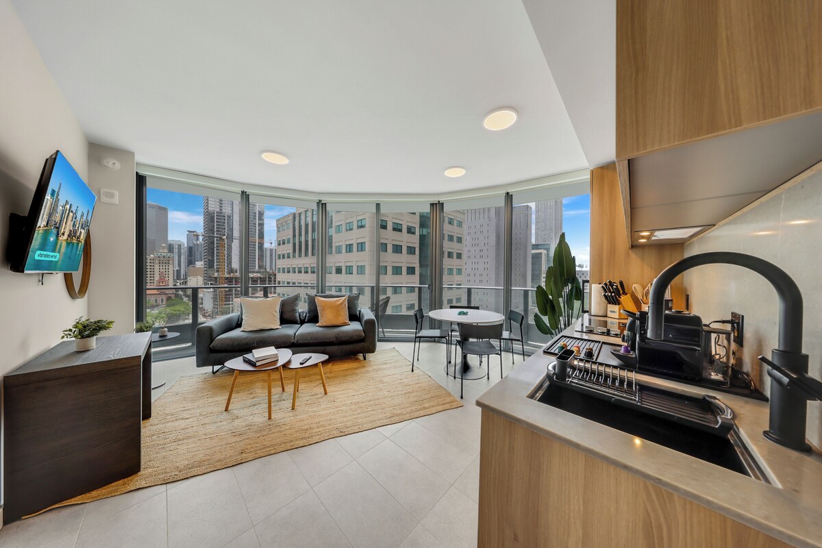 A modern living room with a sofa, coffee table, and a city view through large windows.