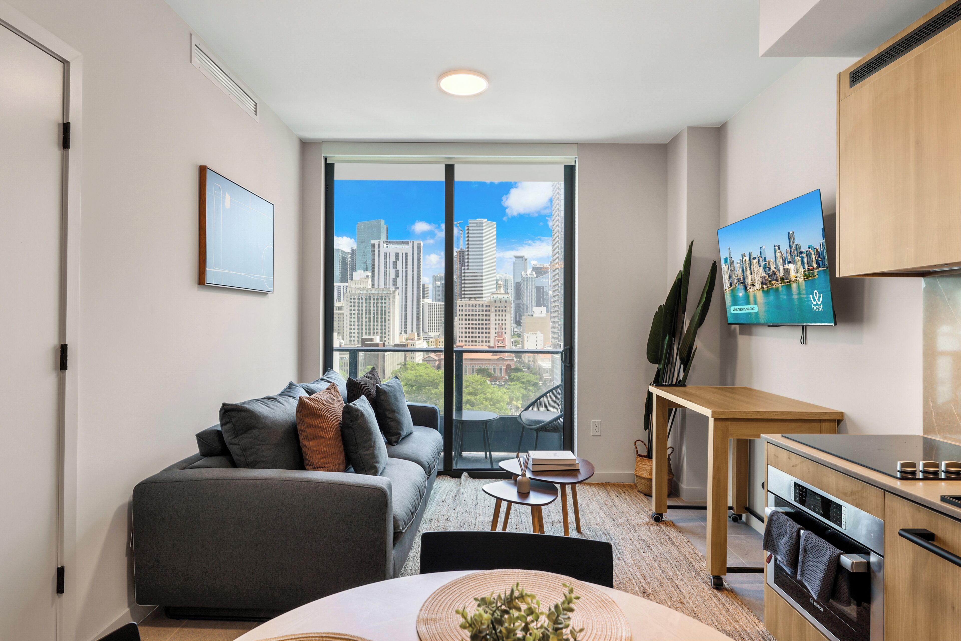 A modern living room with a sofa, a dining table, a kitchen area, and a large window with a city view.