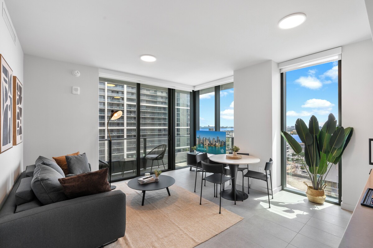 A modern living room with a sofa, coffee table, and chairs, a dining area with a table and chairs, large windows with a city view, and a potted plant.