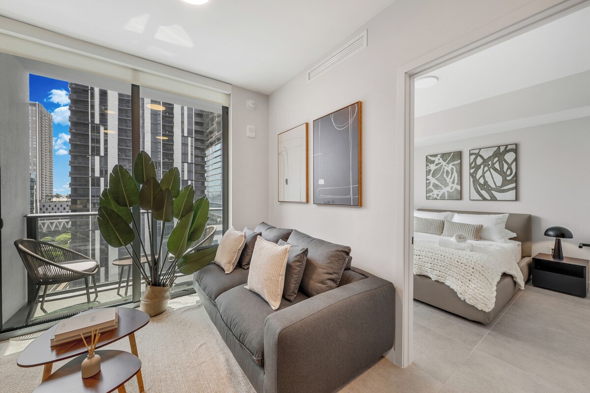 A modern living room with a grey sofa, a coffee table, a potted plant, and a balcony with a view of tall buildings.