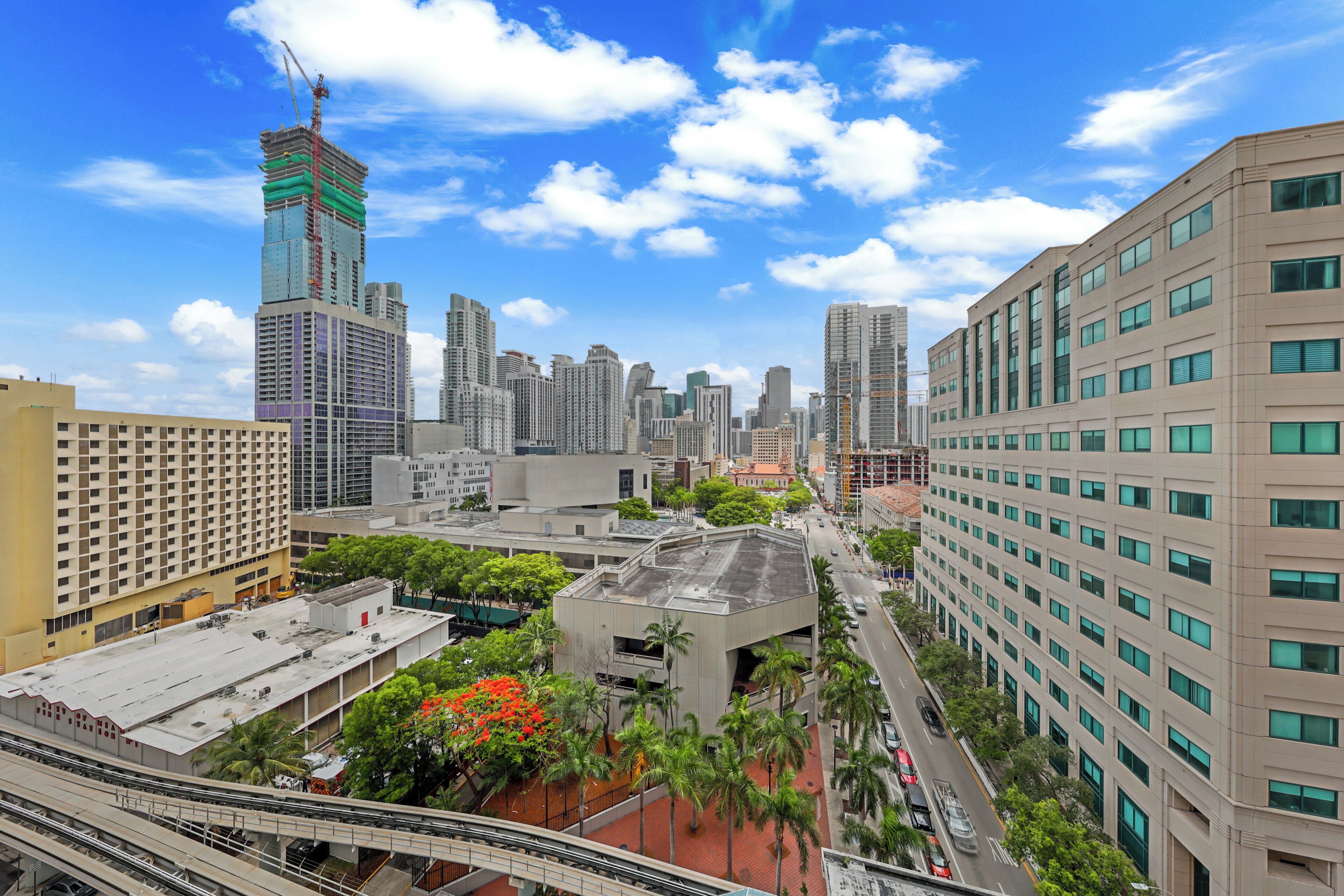 A cityscape with high-rise buildings, a prominent skyscraper under construction, and a pedestrian walkway with palm trees.