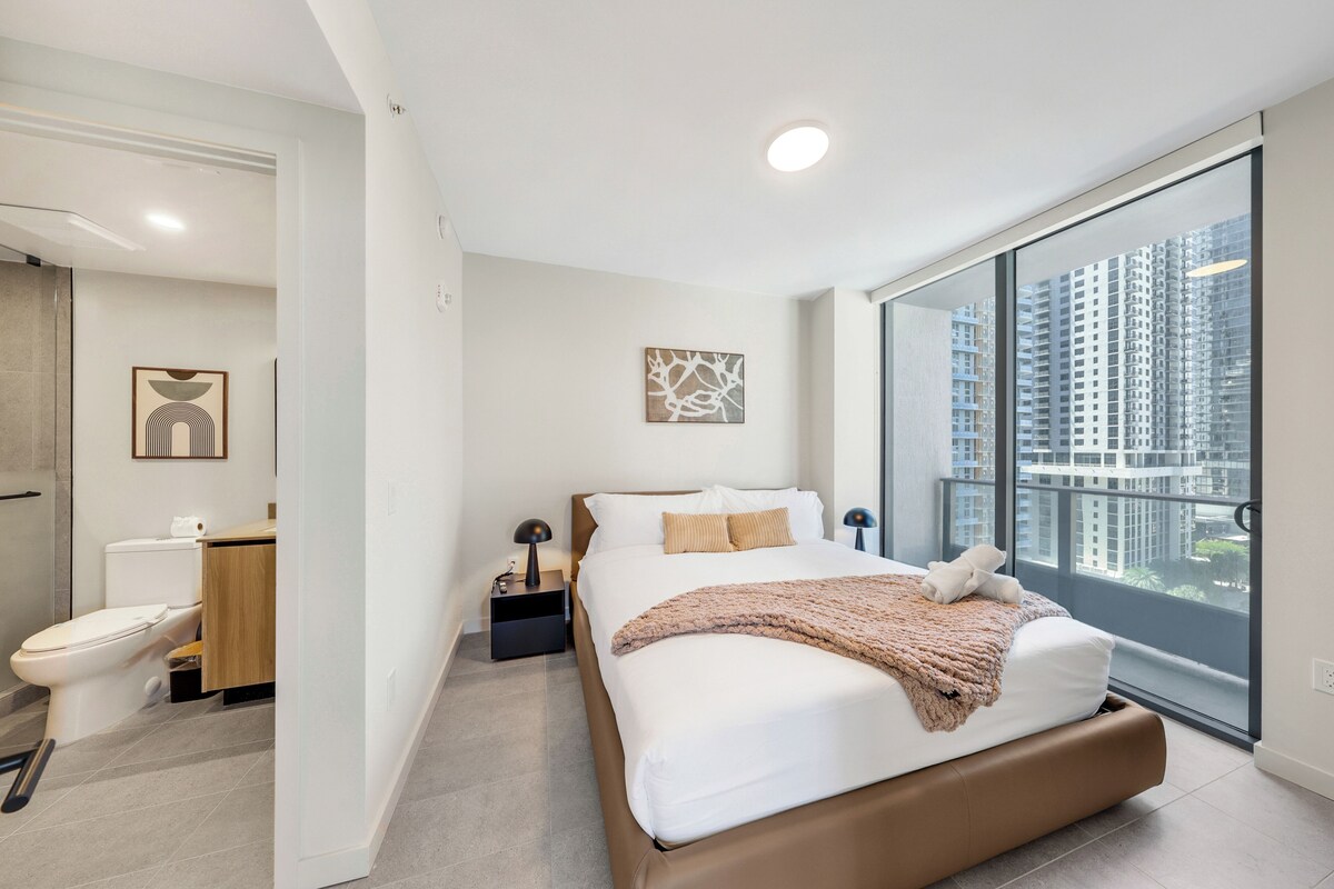 A modern bedroom with a large bed, a nightstand, a bedside table, a window with a city view, and a bathroom with a toilet and sink.