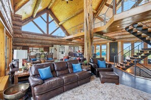 A spacious living area with a high ceiling, wooden beams, a large brown leather sofa, and a staircase.