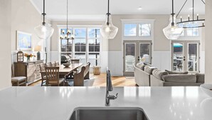 A modern kitchen with a central island, a dining area with wooden chairs, and a living room with a sofa and a view of the outdoors. - Fairview Lake Haven | New Waterfront Home | Private Side | 6BR/4BA | Boathouse & Dock (Mineral)