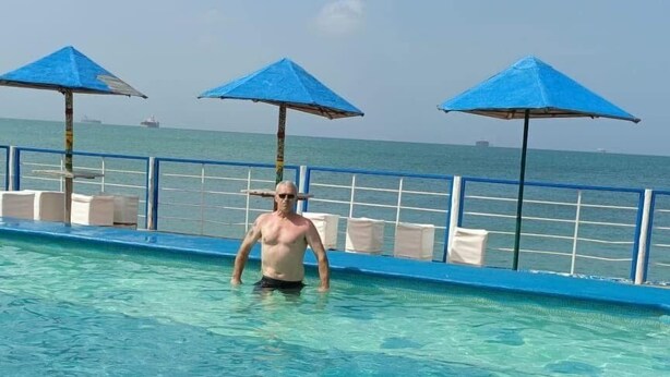 En man i en swimmingpool med klart vatten, blå parasoller och ett räcke.