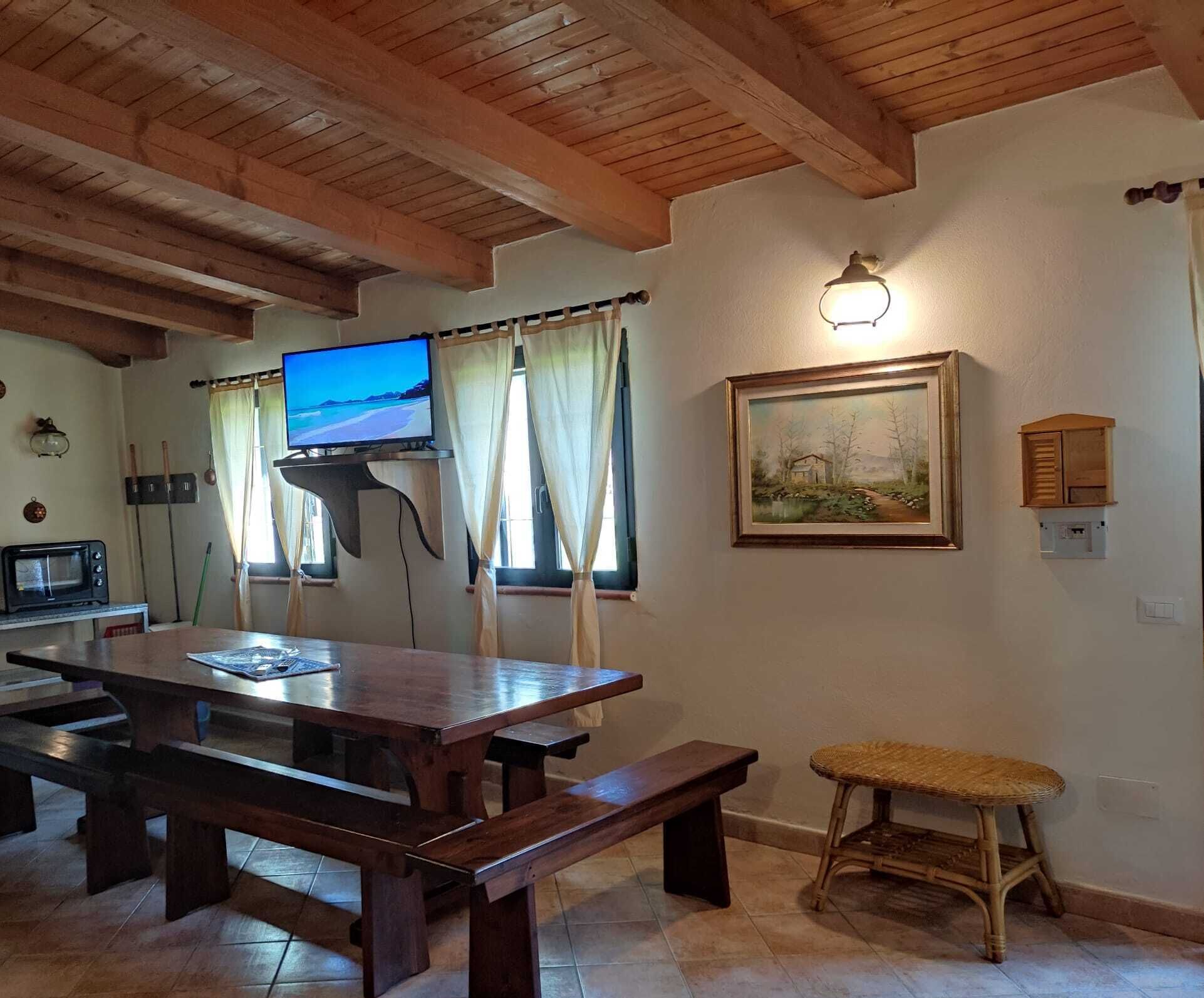 Ein Holztisch mit Bänken, ein Fernseher auf einem Schrank, ein Bild an der Wand und ein Korbstuhl.