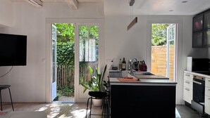 Ett modernt kök med en köksö i mitten, en matplats med bord och stolar, en tv och utsikt över en trädgård genom den öppna dörren.