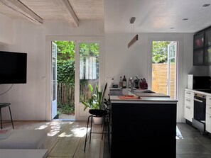 Eine moderne Küche mit einer zentralen Kochinsel, einem Essbereich mit Tisch und Stühlen, einem Fernseher und Blick auf einen Garten durch die offene Tür.